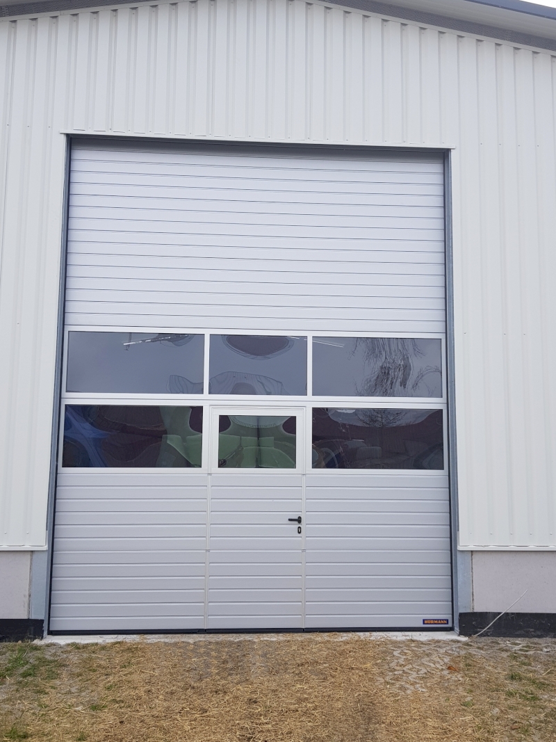 SPU Industrietor, Oberfläche Stucco RAL 9006 Weißaluminium. Mit 2 Alurahmen. Verglasung Kunststoff-Doppelscheibe, klar. Schlupftür mit Panik Drückergarnitur.